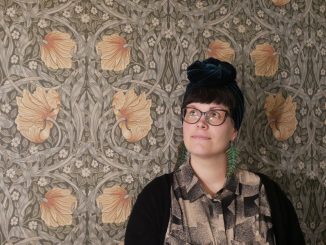 Kuvassa Joanna Heinonen seisoo kukkakuvioisen seinän edessä. Hän katsoo yläviistoon vasemmalle ja hymyilee.